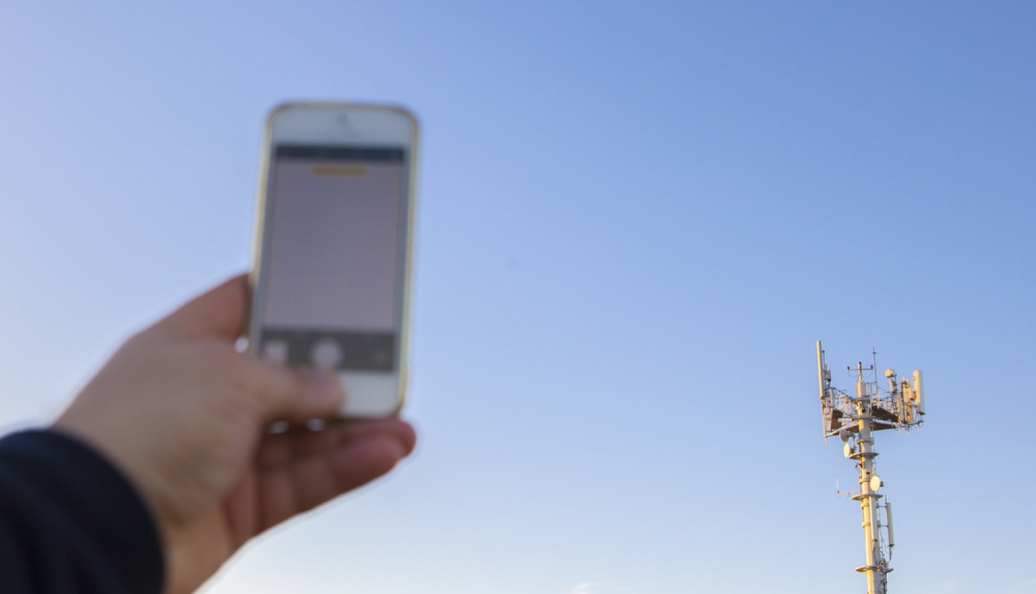 Cilvēks tur rokā mobilo telefonu, fonā sakaru tornis