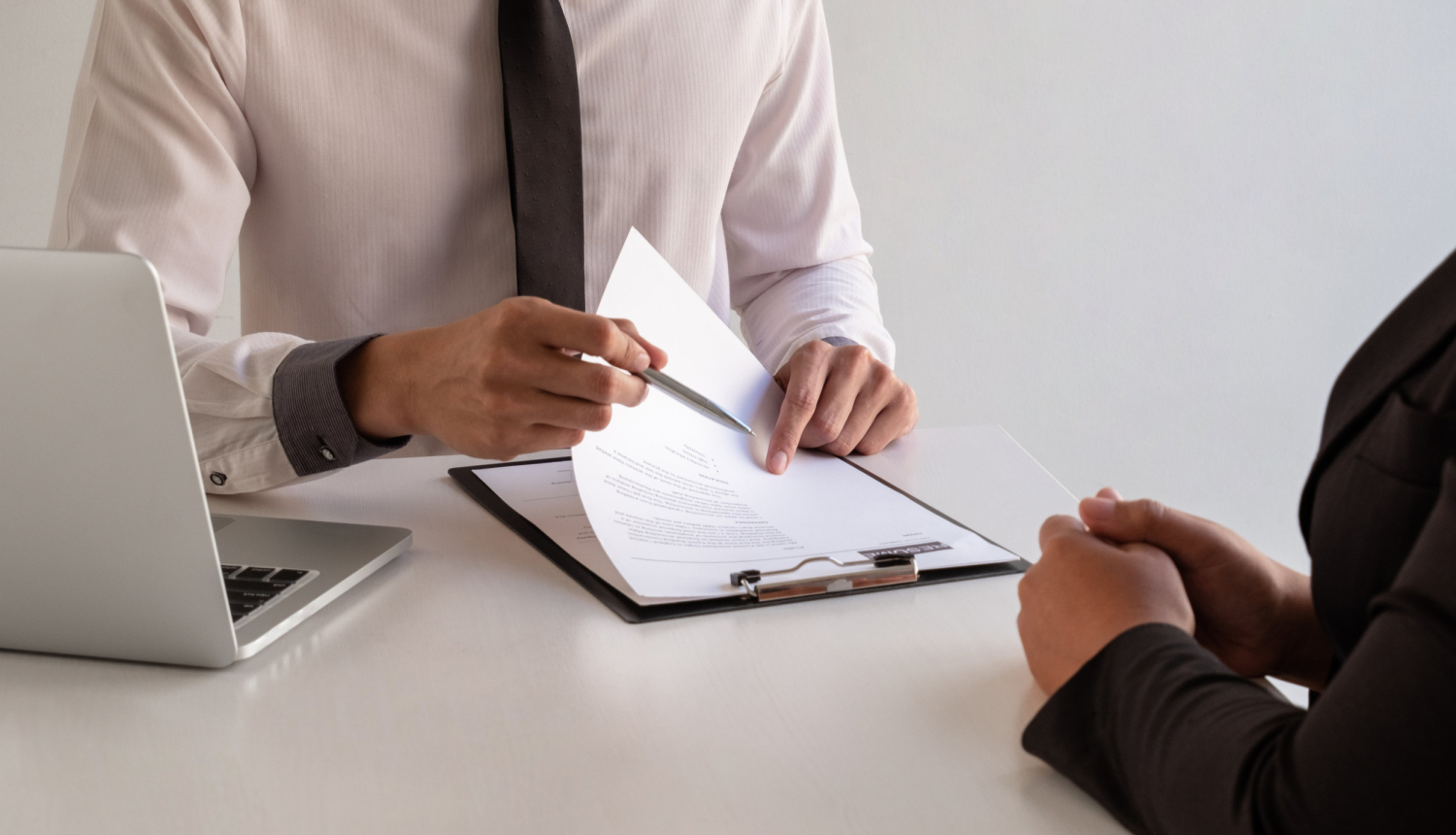 Foto: 2 cilvēki pie galda, viens tur rokā mapīti, uz galda dators