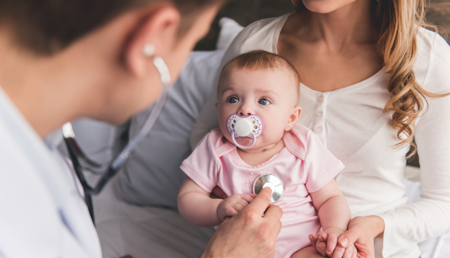 Ģimenes ārsts pārbauda mazuli ar stetoskopu. Mazulis ir rokās mammai. 