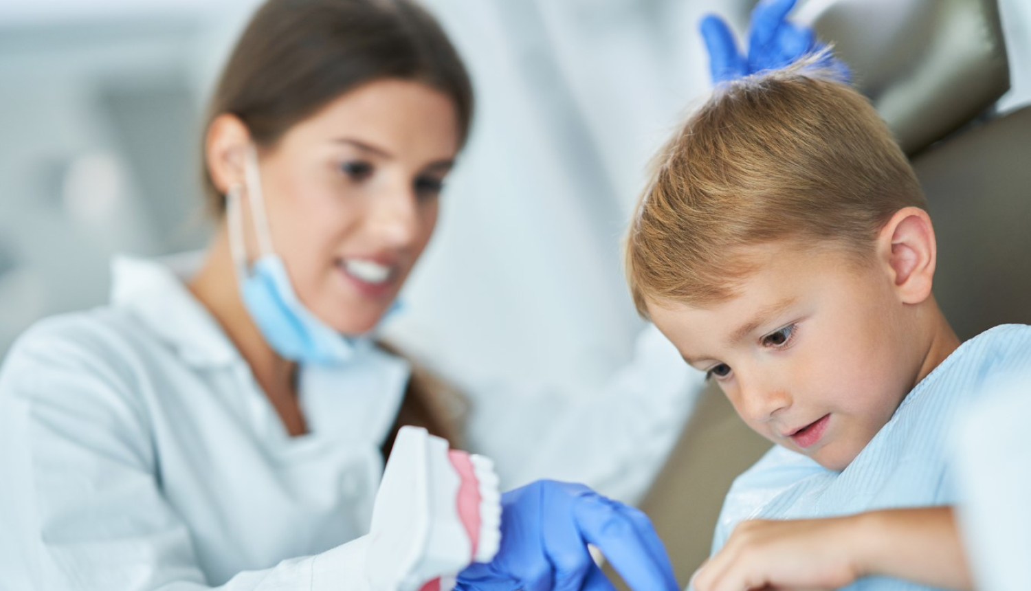 Bērnu zobārsts ar puisi, kurš sēž zobārsta krēslā un rokās ir modelis ar zobiem.