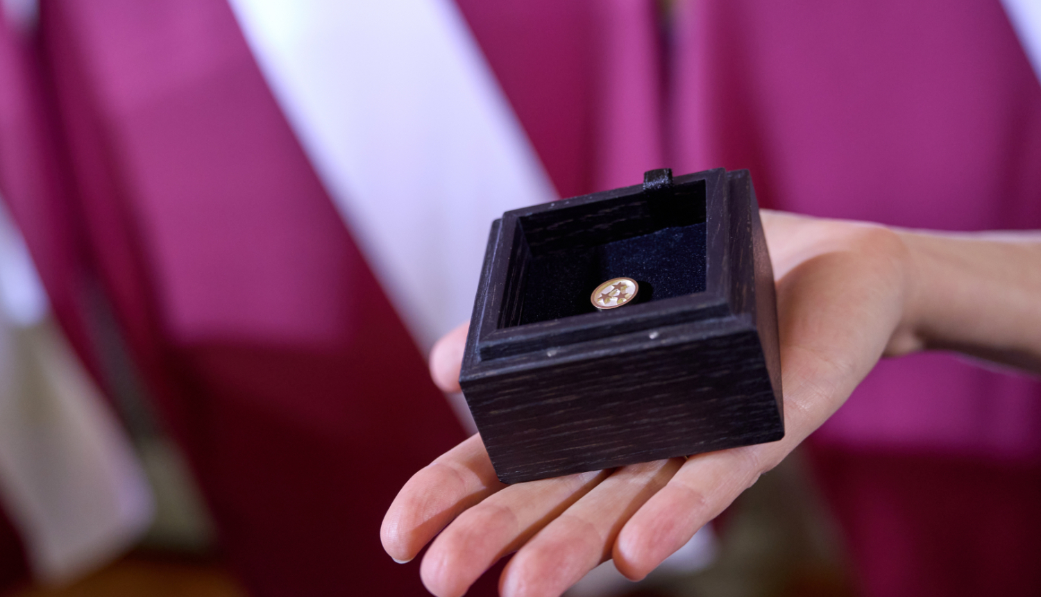 Uz Latvijas karoga fona plaukstā novietota koka kastīte, kurā ievietota Ministru kabineta balvas krūšu nozīmīte