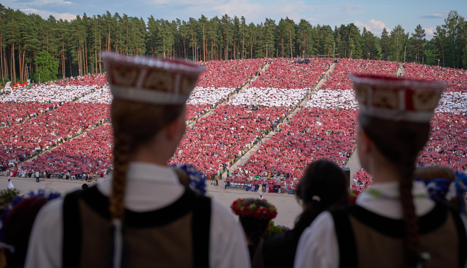 Priekšplānā meiteņu silueti latviešu tautastērpos, tālāk skats uz Mežaparka estrādes skatītāju zonu, kurā skatītāji izveidojuši Latvijas karogu, paceļot gaisā krāsainas papīra lapas