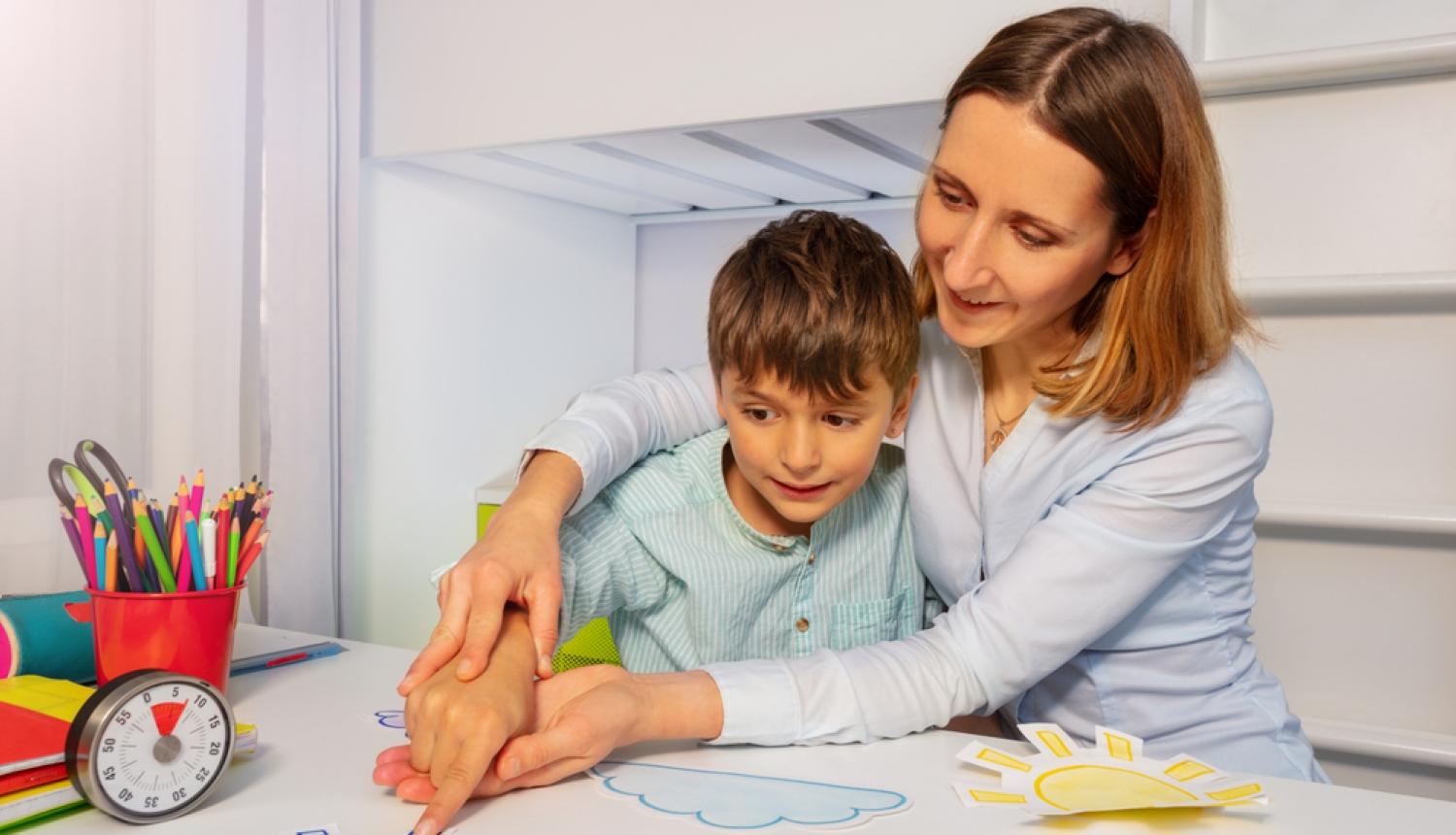 Mamma ar dēlu, kuram ir autisms. Uz galda krāsainas rotaļas. Mamma tur dēla roku 