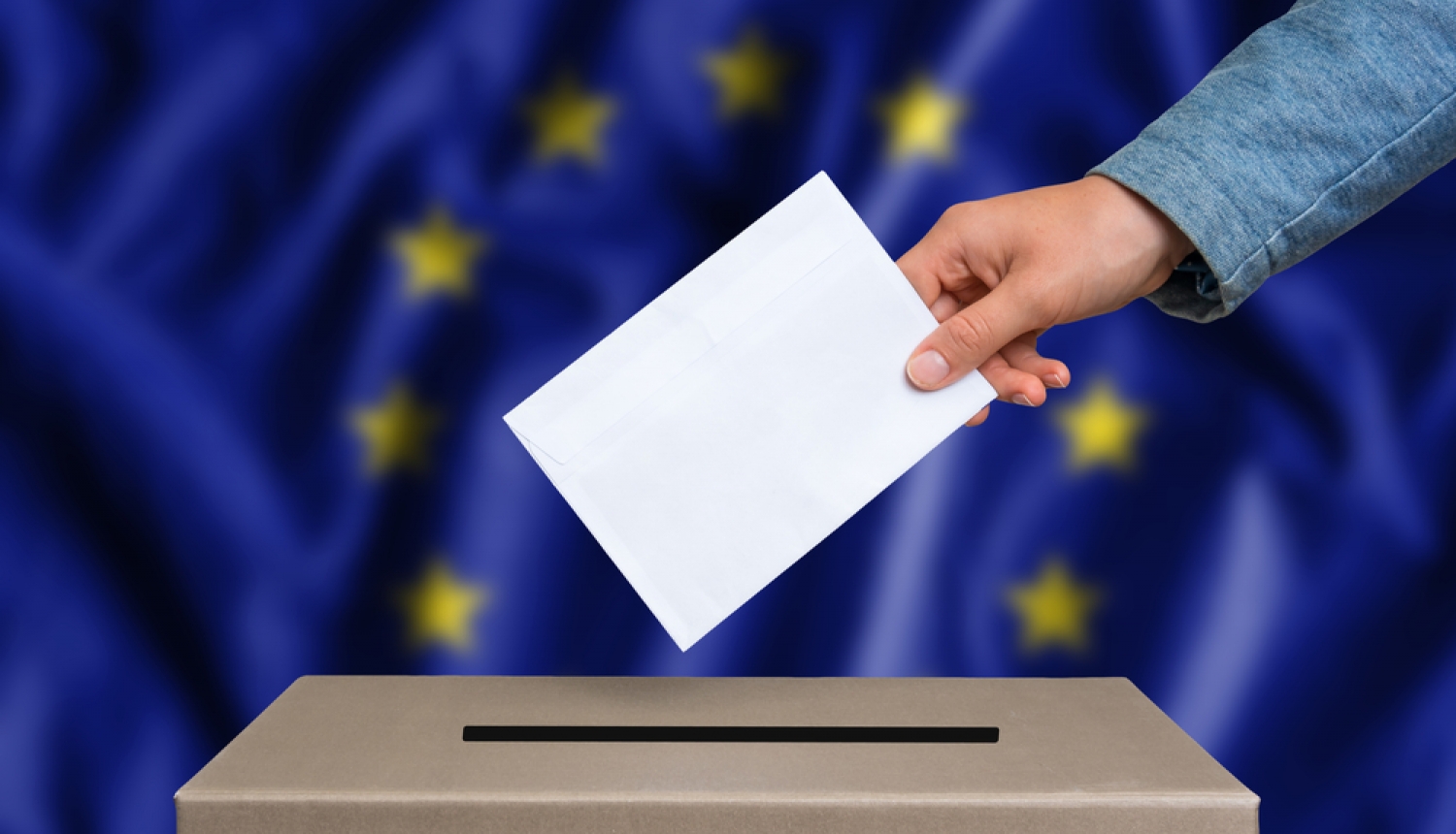 Eiropas Parlamenta vēlēšanas. Roka met kastē aploksni. Fonā Eiropas Savienības karogs. 