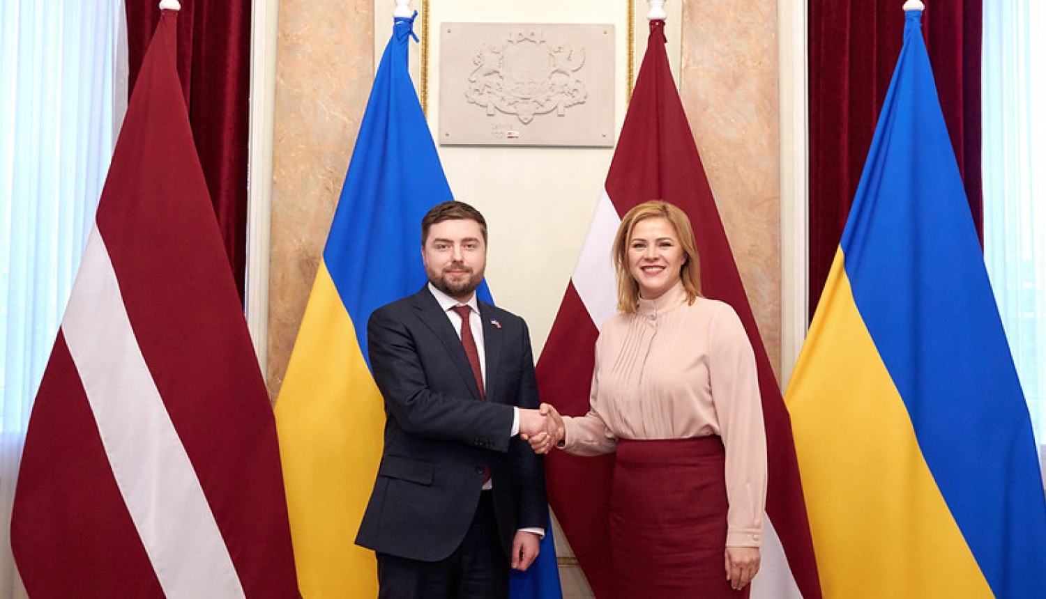 Ministru prezidente Evika Siliņa sarokojas ar Ukrainas vēstnieku Latvijā Anatoliju Kutsevolu. Fonā Latvijas un Ukrainas karogi. 
