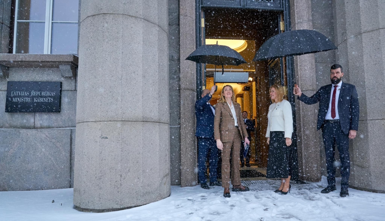 Roberta Metsola un Evika Siliņa pie valdības nama. Snieg sniegs un 2 vīrieši tur virs viņām lietussargus. 