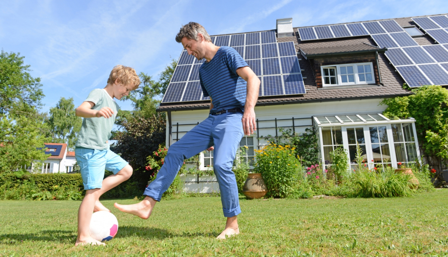 Tētis ar dēlu spēlē futbolu, fonā māja ar saules paneļiem