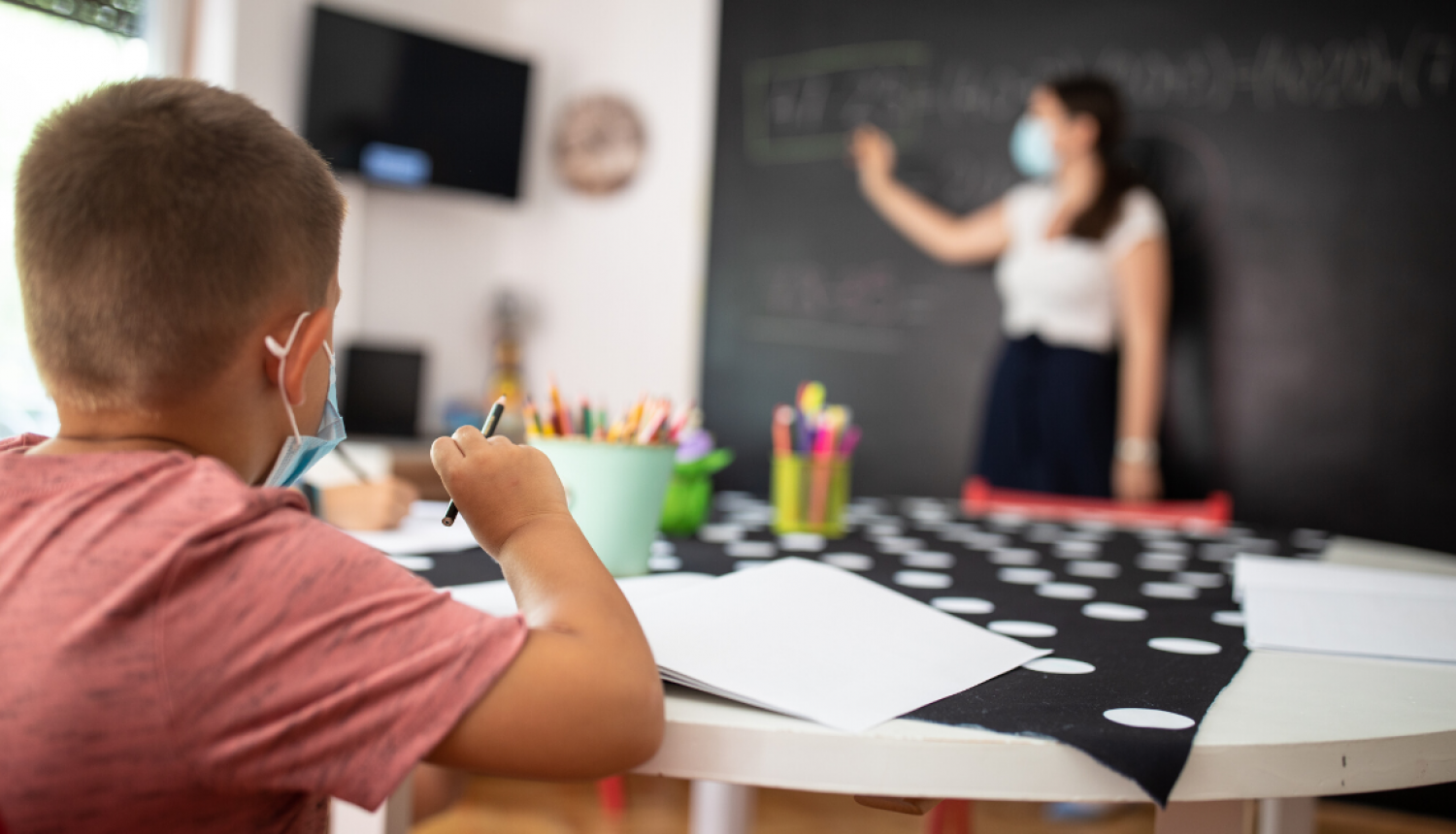 Bērns ar masku skolā pie galda, skatās uz skolotāju un tāfeli