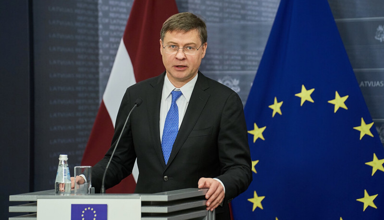 Eiropas Komisijas priekšsēdētājas izpildvietnieks Valdis Dombrovskis preses konferenču telpā. Fonā Latvijas un Eiropas Savienības karogi.